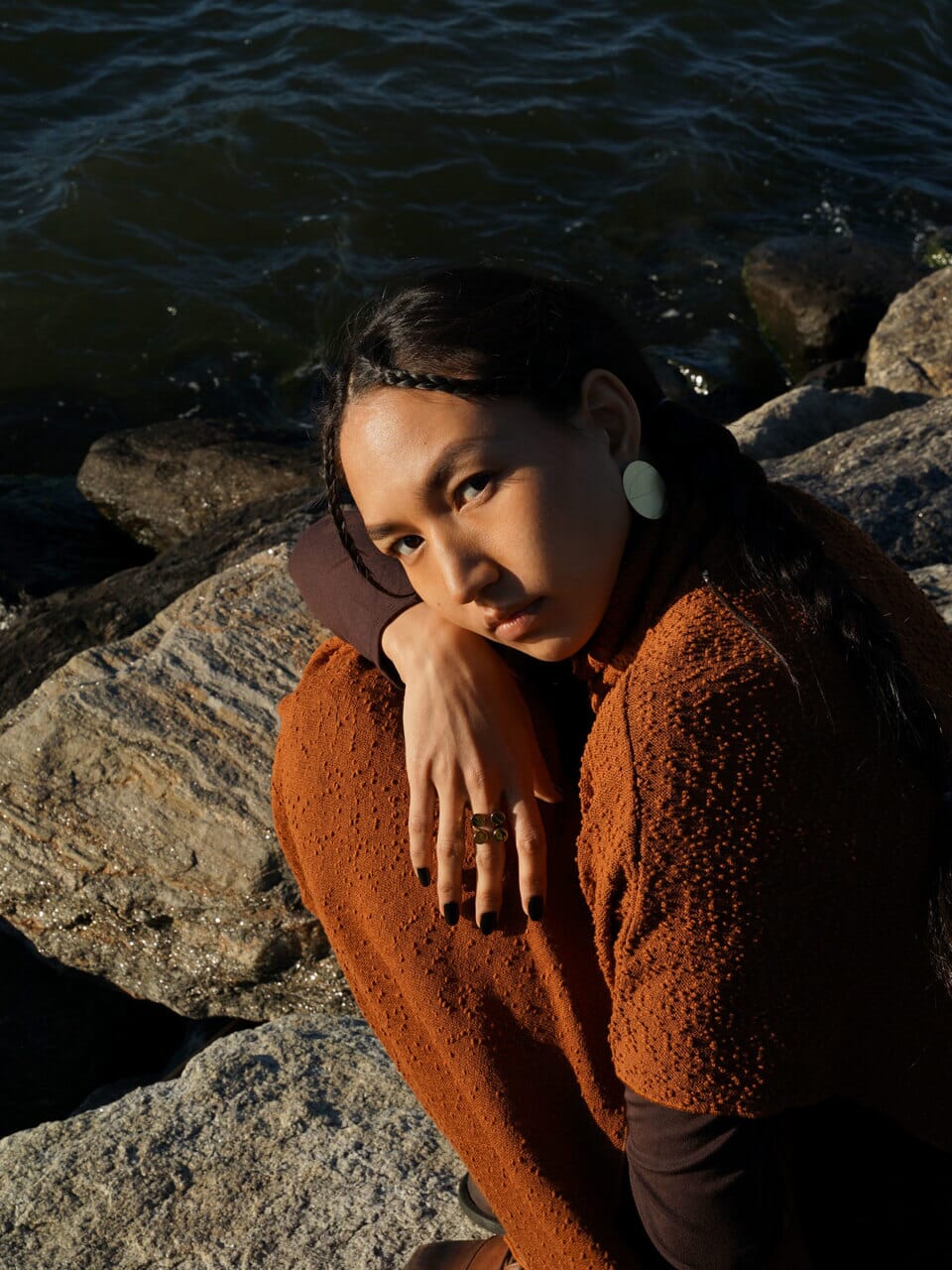 Boho woman sitting on rocks by a body of water, wearing handmade jewellery, earrings and ring