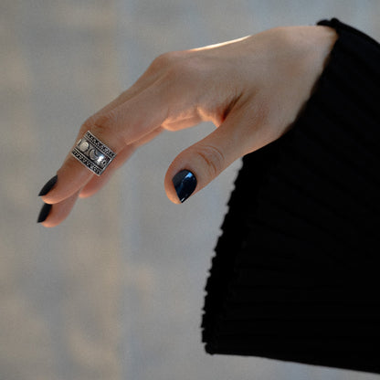Hand wearing a silver ring on celestial theme sun moon stars with black nail polish against a neutral background 