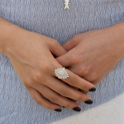 A close-up of a person's hands wearing a large silver ring with a central white pearl and a textured silver band, set against a light blue textured top.