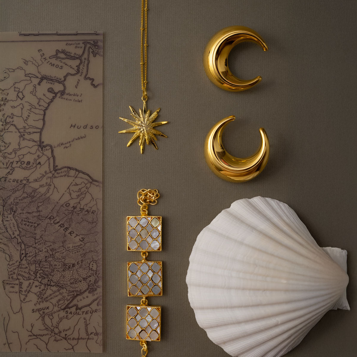 Golden jewelry pieces including a star necklace, mother of pearls square shaped bracelet and crescent moon earrings on a brown surface with a shell and map.