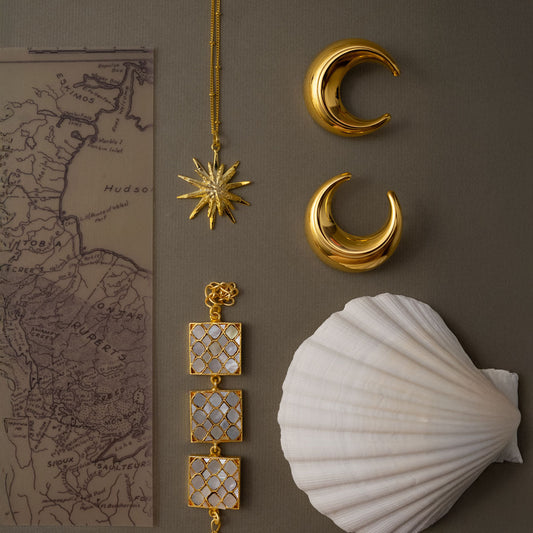 Golden jewelry pieces including a star necklace, mother of pearls square shaped bracelet and crescent moon earrings on a brown surface with a shell and map.
