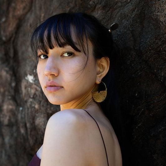 Woman with brass golden hoop earrings with intricate design, leaning against a tree